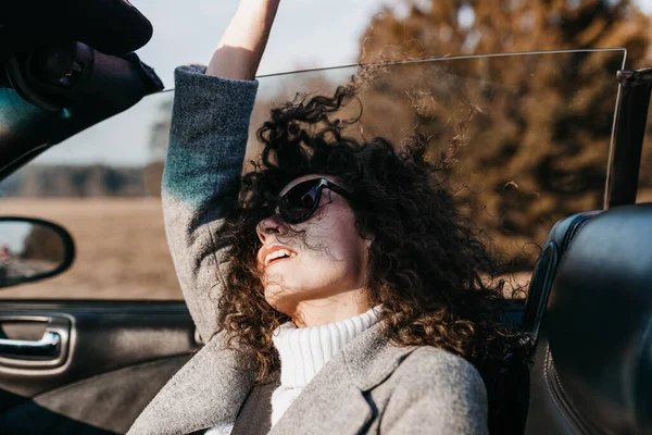 Curly woman photographer in a convertible car rides on a sunny day on the road and rejoices in sunglasses