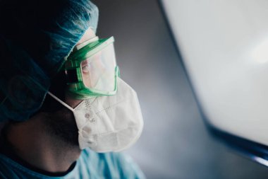 Koruyucu üniformalı bir adam ve epidemiyoloji uzmanı gözlüklü bir adam geceleri hastanede ekrana bakıyor.