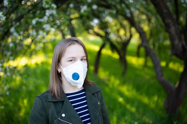 young woman in a medical mask with a valve in a summer park with flowering trees.