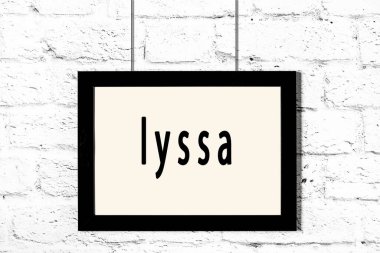 Beyaz tuğla duvarda asılı siyah çerçeve ve Lyssa yazısı.