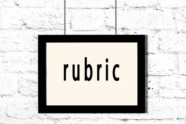 Black frame hanging on white brick wall with inscription rubric — Stock Photo, Image