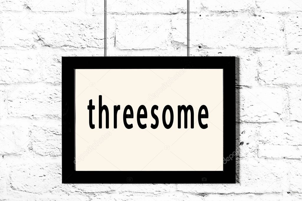 Black frame hanging on white brick wall with inscription threesome