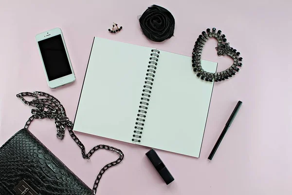 Κάτοψη του τσάντα μαύρη γυναίκα με κενό σημειωματάριο, στυλό, καλλυντικά, αξεσουάρ και Iphone5 σε ροζ φόντο — Φωτογραφία Αρχείου