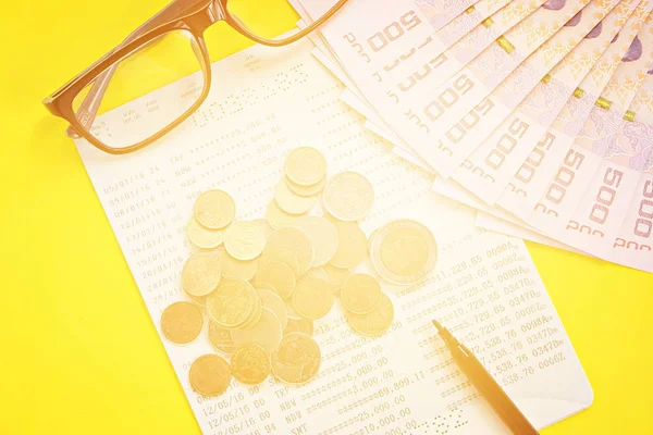Savings account passbook, Thai money, coins, eye glasses and pen on yellow background