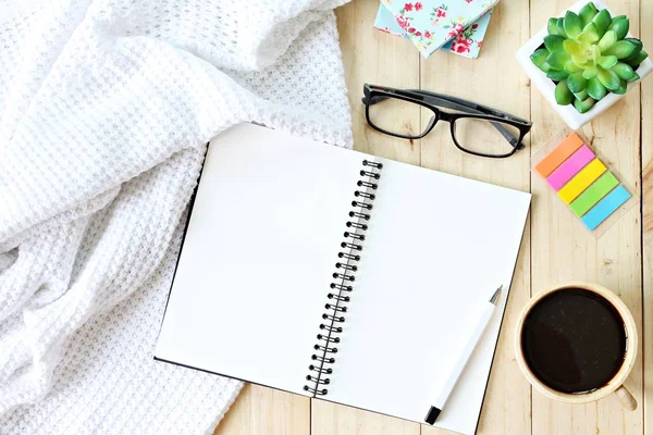 Plat leggen of bovenaanzicht van wit gebreid deken, open lege notebookpapier, koffiekopje en brillen op houten achtergrond — Stockfoto