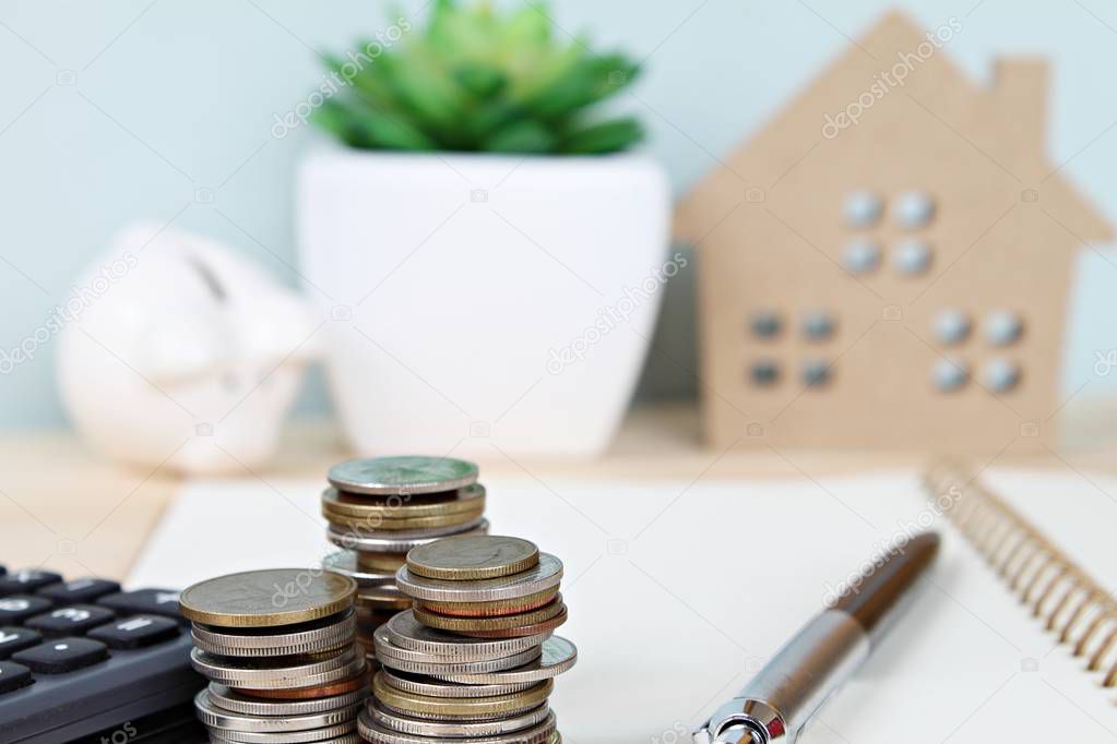Coins stack and calculator in front of wood house model and piggy bank on office desk table