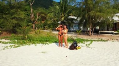 İki genç kız arkadaş kumlu deniz kıyısında durmuş, bir kızın cep telefonunda fotoğraf çekiyor ve izliyorlar. Tropik tatil köyünde dinlenen güzel kadınlar 