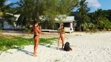 Kumlu deniz kıyısında duran bikinili iki genç kız arkadaş. Bir kız, cep telefonunda meyveli kız arkadaşının fotoğrafını çekiyor. Tropik tatil köyünde dinlenen güzel kadınlar 