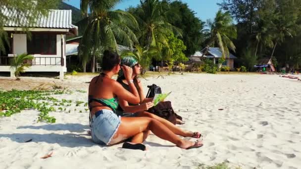 两个年轻的女朋友坐在沙滩上 身边背着行李 看着地图 为将来的旅行和谈话做准备 美丽的女人在热带度假胜地休息 — 图库视频影像