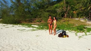 Bikinili iki genç kız, kumlu deniz kıyısında durup cep telefonuyla selfie çekiyorlar. Tropik tatil köyünde dinlenen güzel kadınlar 