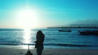 Kadının silueti elinde bavulla sahilde duruyor ve gün batımında akıllı telefon kullanıyor.