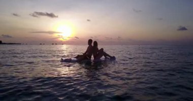 Genç adam ve kadın gün batımında deniz suyunda sörf tahtasında yüzerler. Tropik bir tatil köyünde dinlenen güzel bir çift.   
