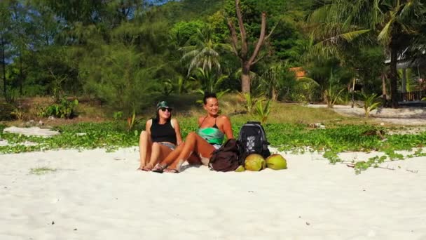 两个年轻的女朋友坐在沙滩上 身边背着行李 在聊天 美丽的女人在热带度假胜地休息 — 图库视频影像