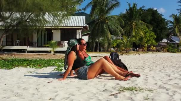 隣の袋を持って砂浜に座って話をしている2人の少女の友人 熱帯リゾートにいる美しい女性 — ストック動画