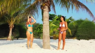 Bikinili iki genç kız arkadaş kumlu deniz kıyısında palmiye ağacının altında güneşleniyor ve konuşuyor. Tropik tatil köyünde dinlenen güzel kadınlar    