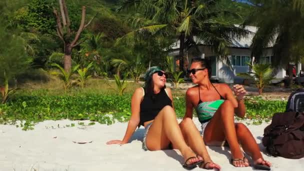两个年轻的女朋友坐在沙滩上 身边背着行李 在聊天 一个女孩用手指头指向某处 美丽的女人在热带度假胜地休息 — 图库视频影像