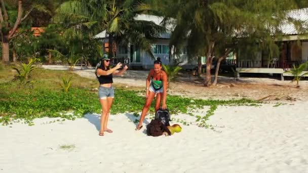 Két fiatal lány barát áll a homokos tengerparton, zacskókkal a mellettük. Egy lány tájképet készít mobilon. Gyönyörű nők pihennek trópusi üdülőhelyen  