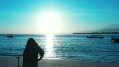 Kadının silueti elinde bavulla sahilde duruyor ve gün batımında akıllı telefon kullanıyor.