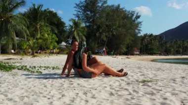 Kumlu deniz kıyısında bavulları yanında oturan ve konuşan iki genç kız arkadaş. Tropik tatil köyünde dinlenen güzel kadınlar   