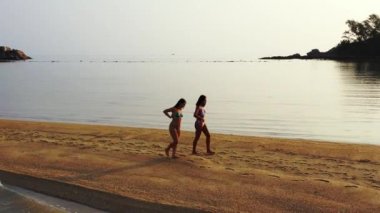 Gün batımında deniz kıyısında yürüyen bikinili iki genç kız arkadaş. Tropik tatil köyünde dinlenen güzel kadınlar      