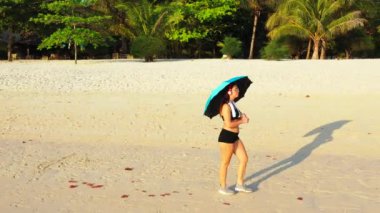 Siyah bikinili muhteşem bir kadın müzik dinliyor ve kumsalda yürürken şemsiye tutuyor. 