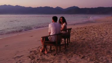 Masanın yanındaki sandalyelerde oturan romantik çift tropikal plajda günbatımının tadını çıkarıyor. 