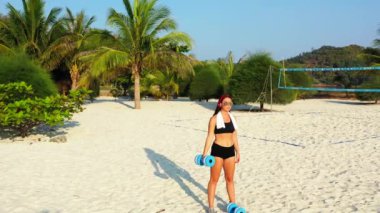 Kadın sporcu kulaklıkla müzik dinliyor ve adanın sahilinde dambıllarla egzersiz yapıyor. 