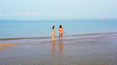 Bikinili iki genç kız deniz kıyısında suyun üzerinde dikilip sohbet ediyorlar. Tropik tatil köyünde dinlenen güzel kadınlar     