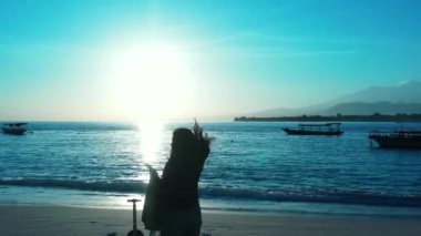 Kadının silueti elinde bavulla sahilde duruyor ve gün batımında akıllı telefon kullanıyor.