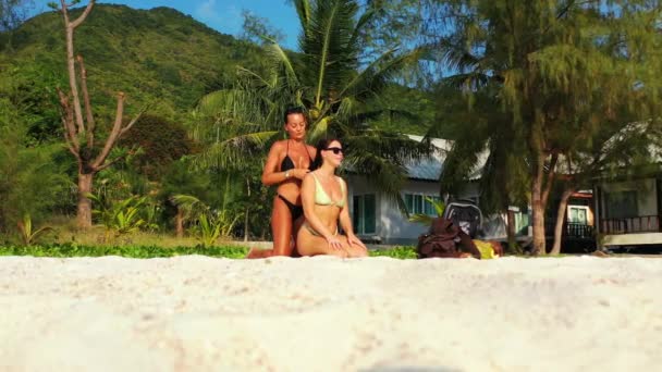 两个年轻的女朋友坐在沙滩上晒日光浴 一个女孩在给她的女朋友做头发 美丽的女人在热带度假胜地休息 — 图库视频影像