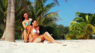 Bikinili iki genç kız arkadaş kumlu deniz kıyısında palmiye ağacının altında güneşleniyor. Bir kız arkadaşını tarak ile tarıyor. Tropik tatil köyünde dinlenen güzel kadınlar    
