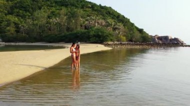 Bikinili iki genç kız deniz kıyısında dikilip akıllı telefondan bir şeyler izliyorlar. Tropik tatil beldesinde güzel kadınlar     