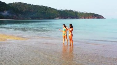 Deniz kıyısında suda duran bikinili iki genç kız arkadaş. Cep telefonunda fotoğraf çeken bir kız. Tropik tatil köyünde dinlenen güzel kadınlar    