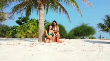 Bikinili iki genç kız arkadaş kumlu deniz kıyısında palmiye ağacının altında güneşleniyor. Bir kız arkadaşının üzerine güneş kremi sürüyor. Tropik tatil köyünde dinlenen güzel kadınlar   
