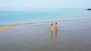 Deniz suyunda yürüyen bikinili iki genç kız arkadaş. Bir kız cep telefonu kullanıyor. Tropik tatil köyünde dinlenen güzel kadınlar