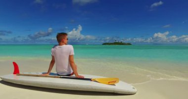 Okyanus kıyısındaki SUP küreğinde oturan genç adam.