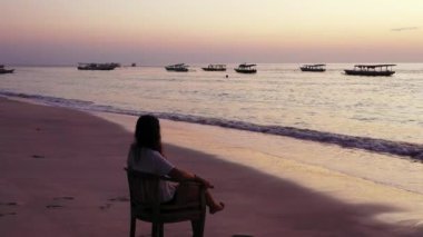 Kumlu sahilde gün batımında sandalyede oturan bir kız.