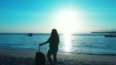 Gün batımında elinde bavulla sahilde duran bir kadın silueti.