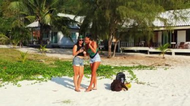 Kumlu deniz kıyısında bavulları yanında duran ve cep telefonundan bir şey izleyen iki genç kız arkadaş. Tropik tatil köyünde dinlenen güzel kadınlar 