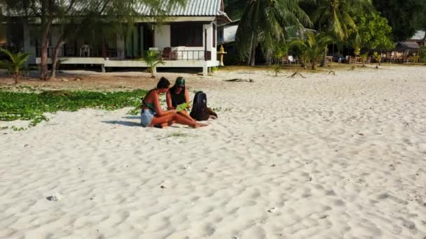 两个年轻的女朋友坐在沙滩上 身边背着行李 看着地图 为将来的旅行和谈话做准备 美丽的女人在热带度假胜地休息 — 图库视频影像