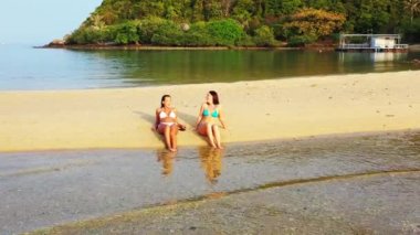 Bikinili iki genç kız deniz kıyısında güneşleniyor ve konuşuyor. Tropik tatil köyünde dinlenen güzel kadınlar    