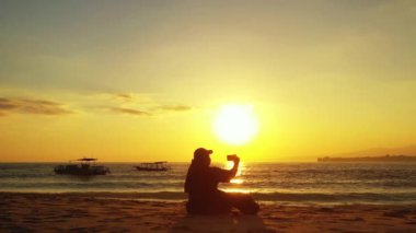 Kadın sahilde oturuyor ve gün batımında fotoğraf çekiyor.