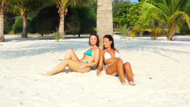 两个穿着比基尼的年轻女友坐在棕榈树下的沙滩海岸上 晒日光浴 聊着天 美丽的女人在热带度假胜地休息 — 图库视频影像