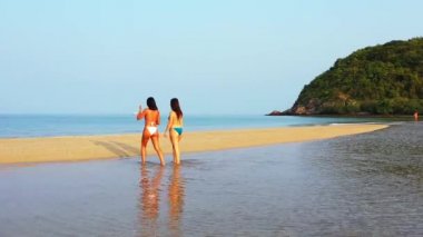 Senkronize bir günde deniz kıyısında yürüyen bikinili iki genç kız arkadaş. Arka manzara Tropik tatil köyünde dinlenen güzel kadınlar    