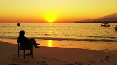 Gün batımında sahilde oturan bir kadın silueti.