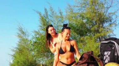 Kumlu deniz kıyısında oturmuş güneşlenen ve konuşan iki genç kız arkadaş. Bir kız arkadaşının üzerine güneş kremi sürüyor. Tropik tatil köyünde dinlenen güzel kadınlar   