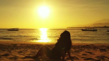 Gün batımında sahilde oturan bir kadın silueti.