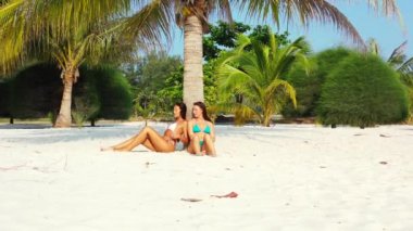 Bikinili iki genç kız arkadaş kumlu deniz kıyısında palmiye ağacının altında güneşleniyor ve konuşuyor. Tropik tatil köyünde dinlenen güzel kadınlar     