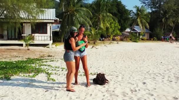 两个年轻的女朋友站在沙滩上 身边背着行李 一边看手机里的东西 美丽的女人在热带度假胜地休息 — 图库视频影像