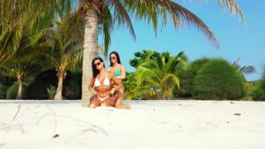 Bikinili iki genç kız arkadaş kumlu deniz kıyısında palmiye ağacının altında güneşleniyor. Bir kız arkadaşının üzerine güneş kremi sürüyor. Tropik tatil köyünde dinlenen güzel kadınlar   